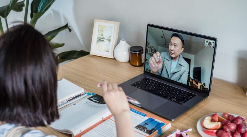 A young woman engaged in an online learning session via laptop surrounded by study materials and snacks on a wooden table.