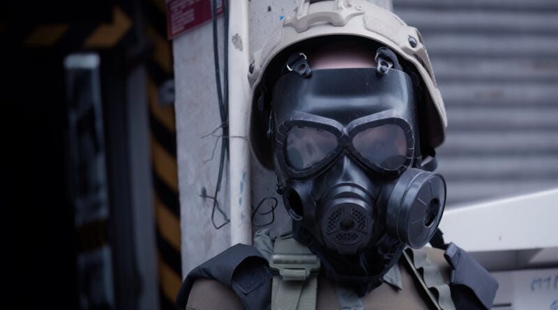 A soldier wearing protective gear and a gas mask standing outdoors against a wall.