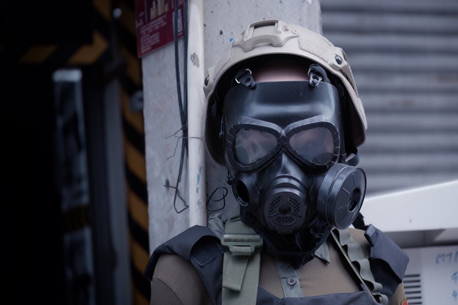 A soldier wearing protective gear and a gas mask standing outdoors against a wall.