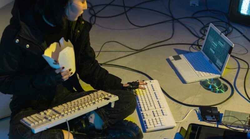 A young woman in a dark room surrounded by computers and cables, eating and typing on keyboards.
