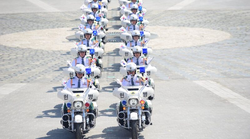 A line of South Korean police officers on motorcycles holding flags in an urban parade.