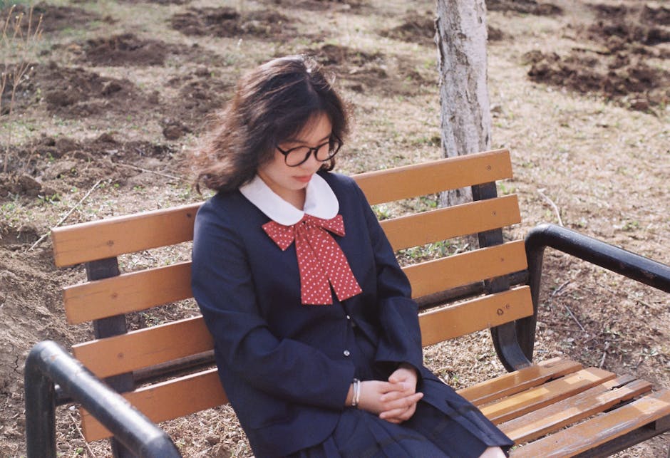 A young woman with glasses sits on a wooden park bench outdoors, showcasing a red bow tie.