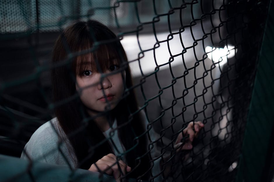 Atmospheric portrait of a teenager behind a chain link fence. Dim lighting creates a dramatic, introspective mood.