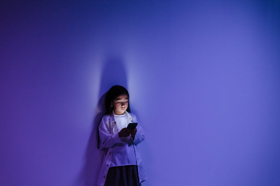 A girl standing against a blue-lit wall engrossed in her smartphone, creating a dramatic silhouette.