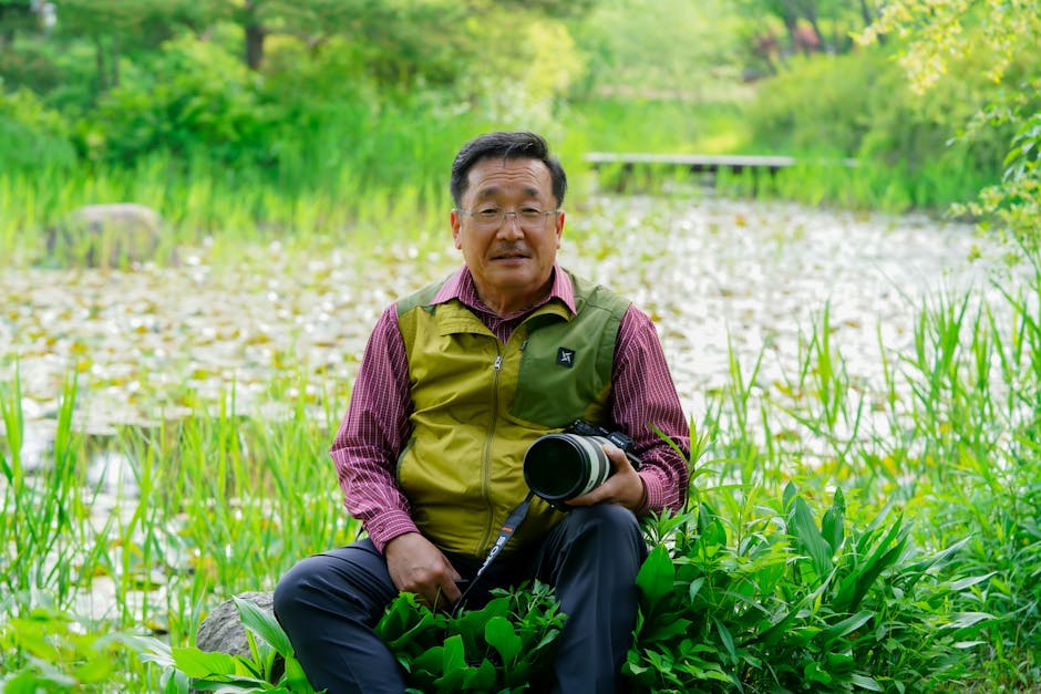 An Asian man sitting by a pond, holding a camera amidst lush greenery.