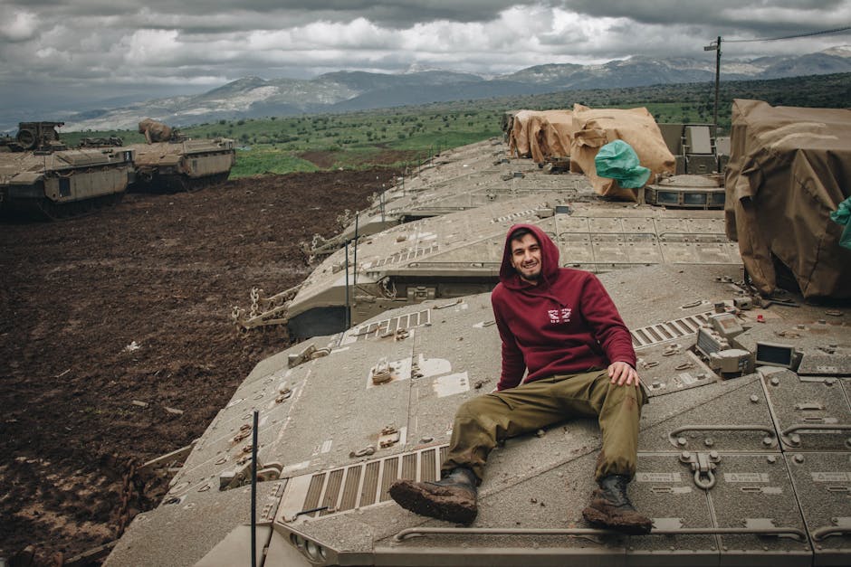 Young man in red hoodie sits on a tank in rural Lebanon, smiling.