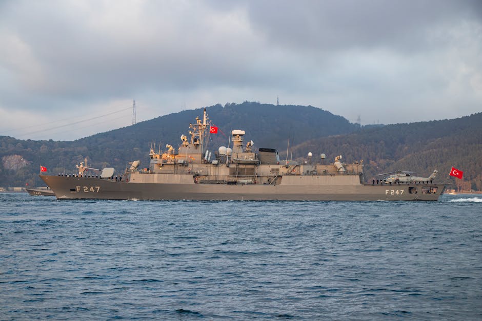 Turkish warship F247 cruising through calm waters with a scenic mountainous backdrop.