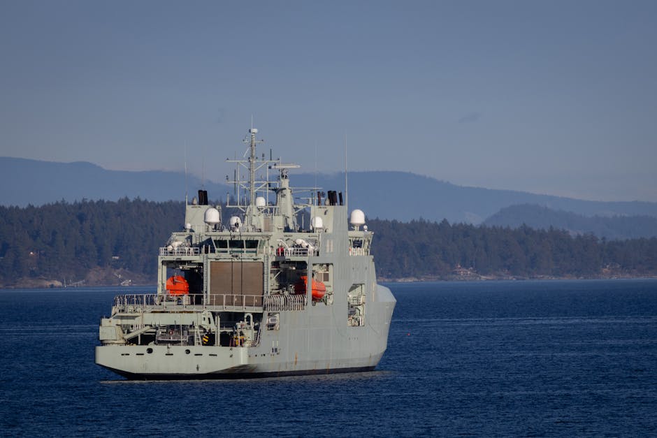 A military warship navigates through open waters with a coastline backdrop on a clear day.