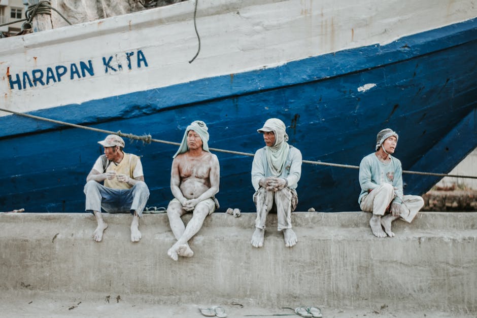 Four dock workers rest on a pier beside a large blue ship, reflecting a moment of pause.