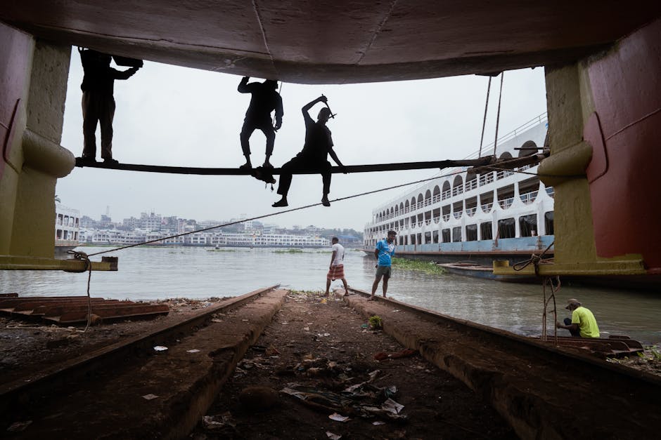 Silhouetted workers engage in maintenance under a docked ship in an urban riverside setting.