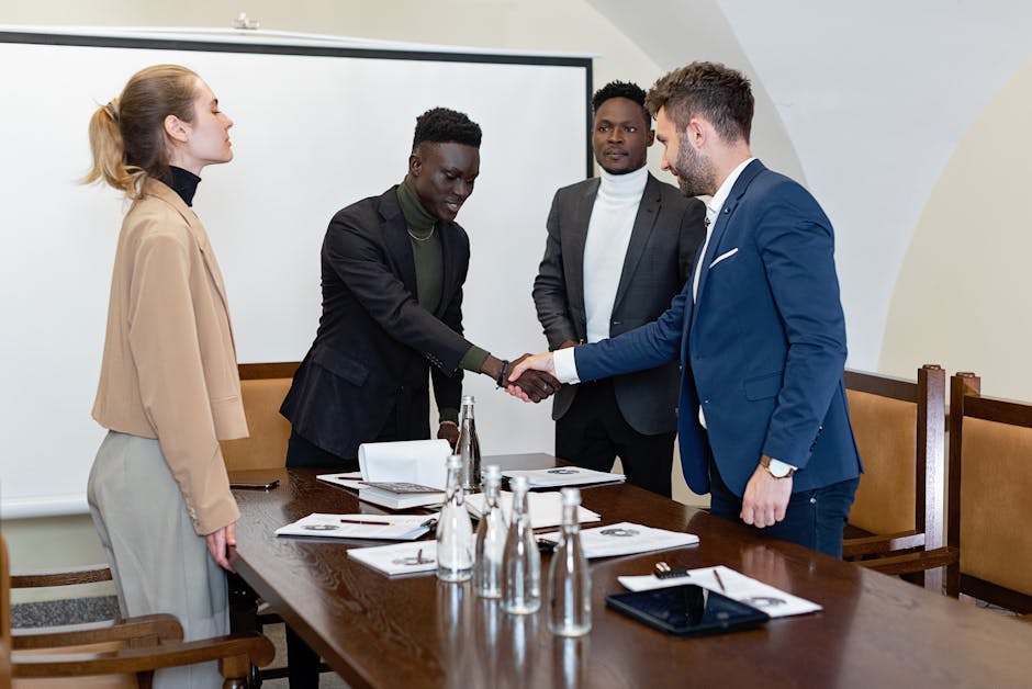 Four professionals in a modern office, two men shaking hands signifying agreement.