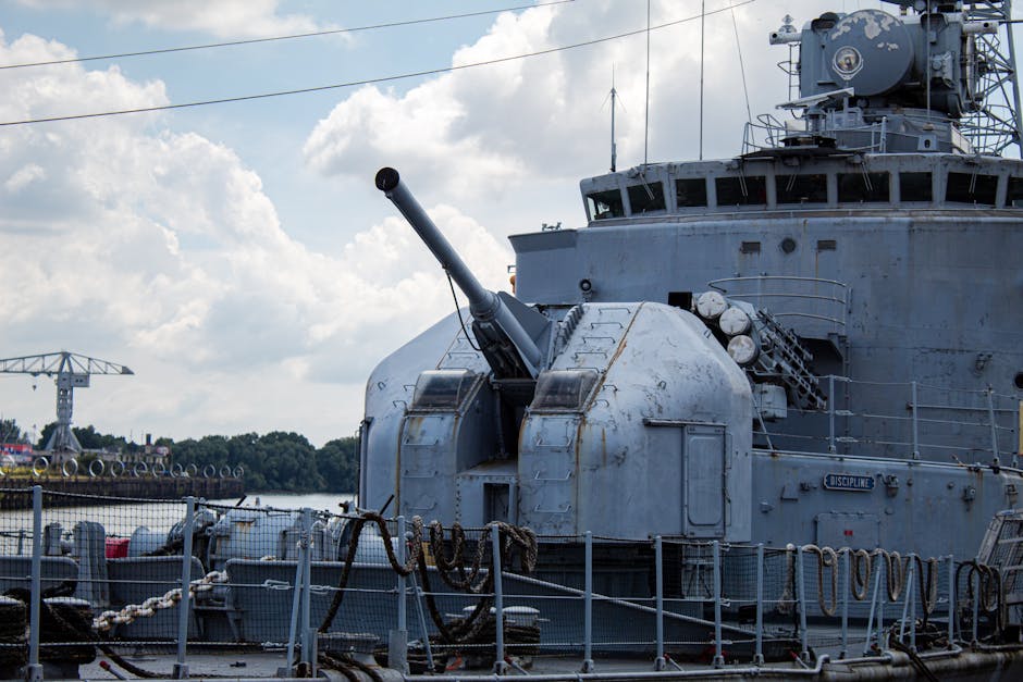 Close-up of a naval warship cannon on deck at a harbor, showcasing its heavy machinery.